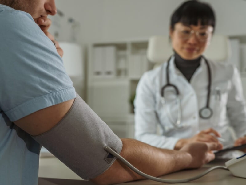 A White man in a light blue t shirt has a blood pressure cuff around his right arm, he has his back to the camera. A young Asian woman in a white doctor's coat with a stethoscope around her beck is taking notes
