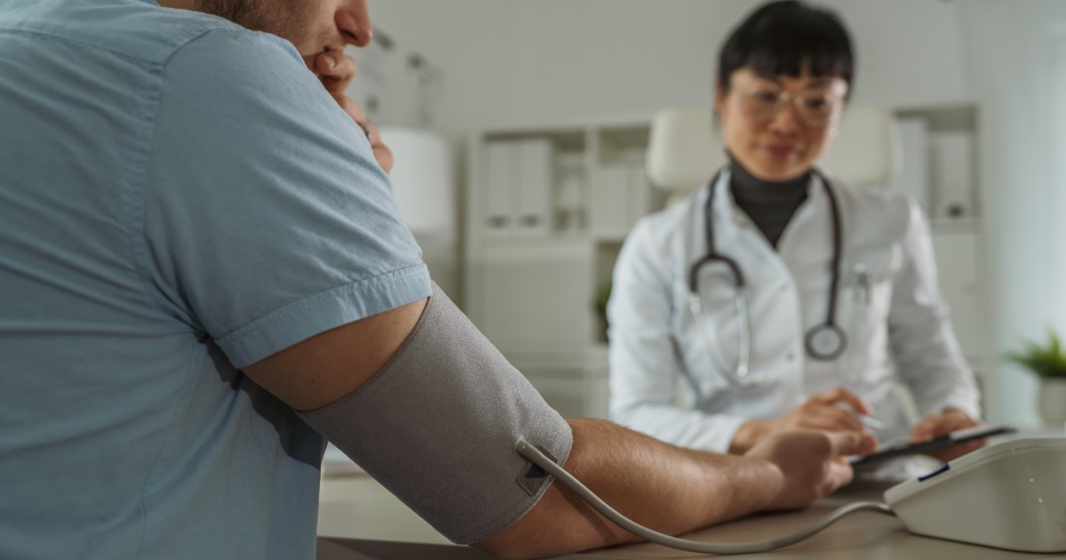 A White man in a light blue t shirt has a blood pressure cuff around his right arm, he has his back to the camera. A young Asian woman in a white doctor's coat with a stethoscope around her beck is taking notes