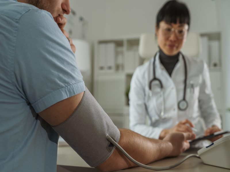 A White man in a light blue t shirt has a blood pressure cuff around his right arm, he has his back to the camera. A young Asian woman in a white doctor's coat with a stethoscope around her beck is taking notes
