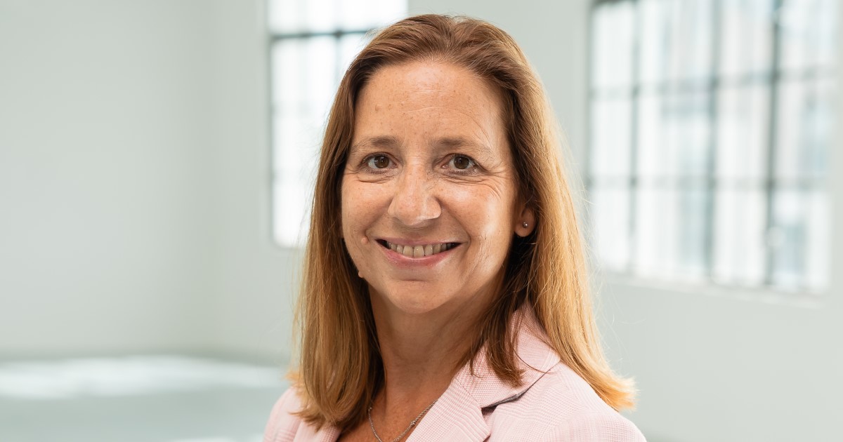 A smiling white red-haired woman stands side on but is turning her head towards the camera. She is wearing a light pink blazer and a black top underneath. She has a red BHF logo pin on her lapel. She stands in an empty white office room. 