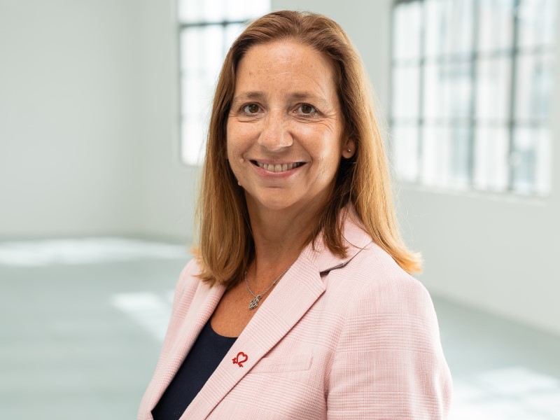 A smiling white red-haired woman stands side on but is turning her head towards the camera. She is wearing a light pink blazer and a black top underneath. She has a red BHF logo pin on her lapel. She stands in an empty white office room. 