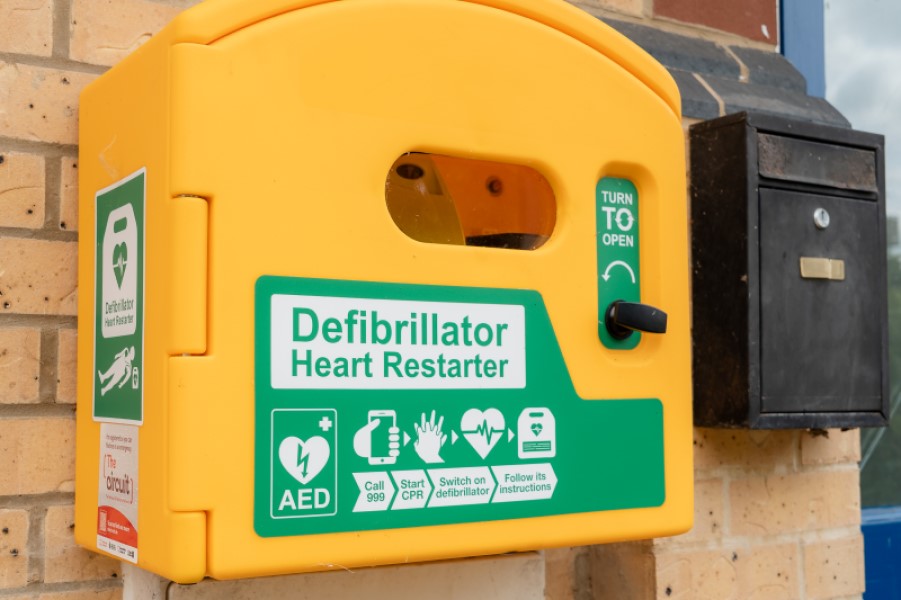 A yellow box on the wall of a grey bricked building. It's a close up shot and in green writing it says Defibrillator Heart Restarter
