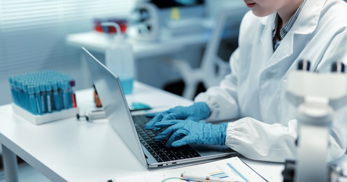 A researcher in a lab coat sits at a lab bench and types with gloved hands on a laptop
