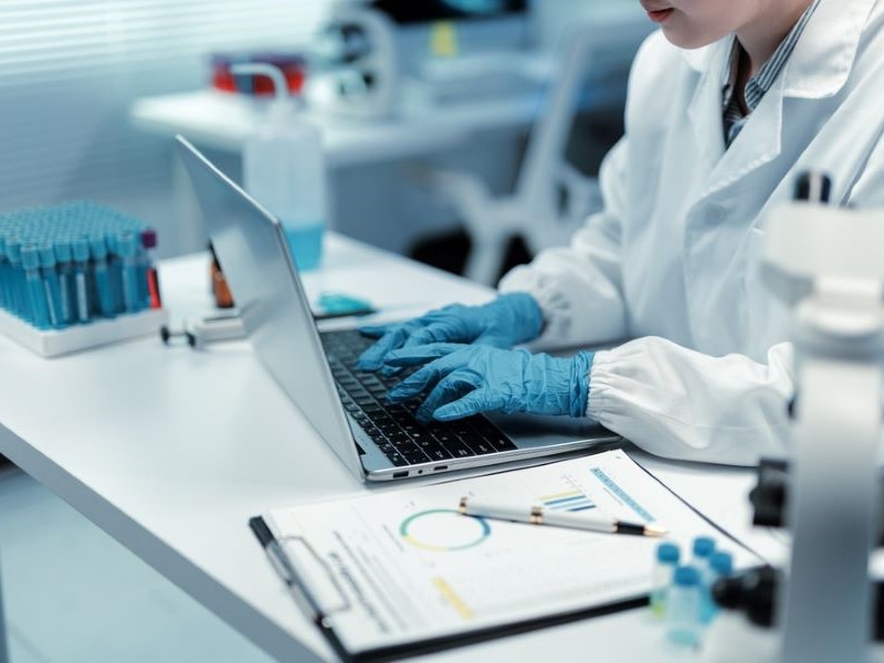 A researcher in a lab coat sits at a lab bench and types with gloved hands on a laptop