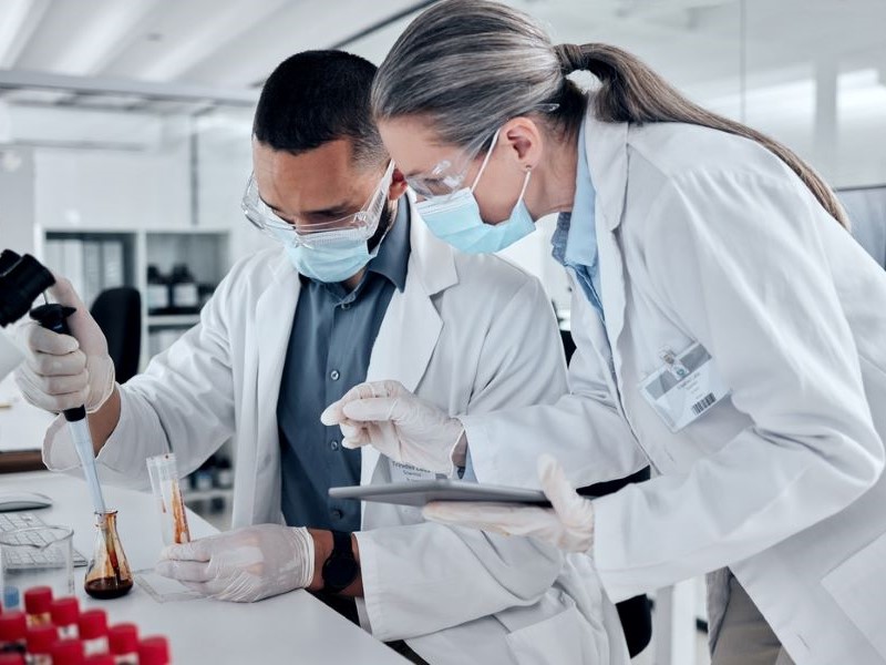 A younger researcher sits at a bench pipetting, whilst an older researcher leans over them and instructs