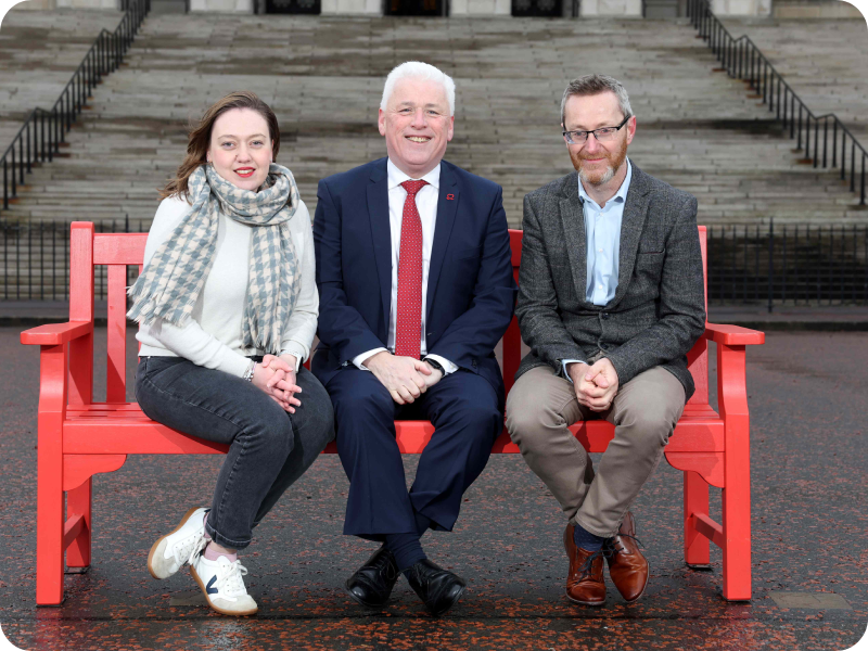 Three white people sit on a red bench in front of Stormont in Northern Ireland