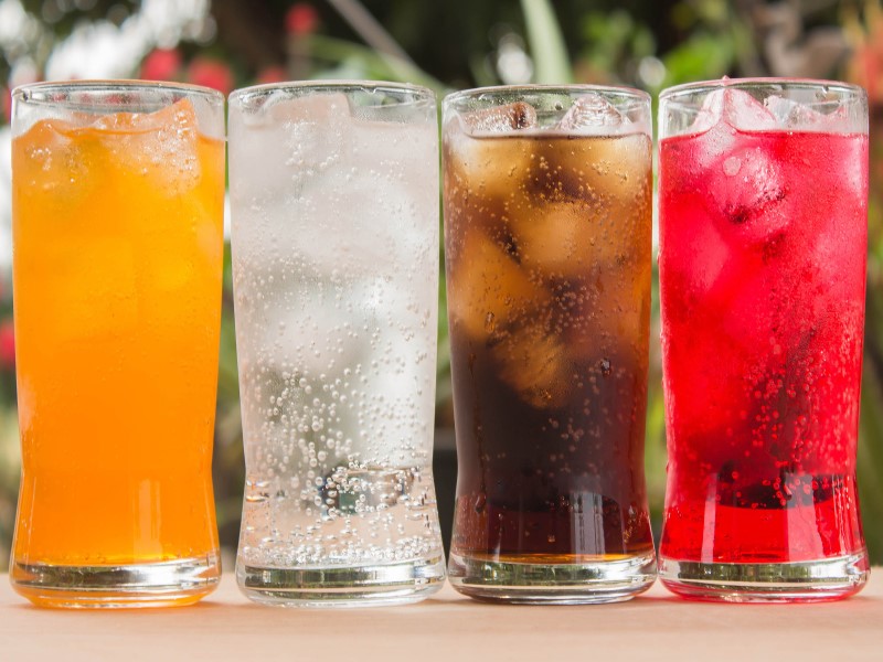 Four fizzy drinks lined up on a wooden table against a garden backdrop. The drinks left to right are: orangeade, lemonade, cola, and a bright red fizzy drink.