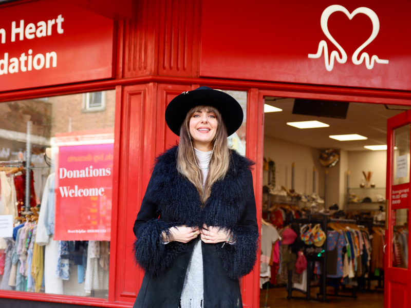 Lauren Cooley wearing a black coat and hat and smiling, standing in front of a British Heart Foundation shop. 