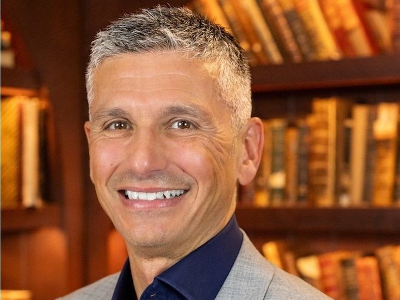 Professor Tim Chico. He is a white man smiling with white/grey hair. Only his face and shoulders show. He is wearing a light grey blazer over a navy blue shirt. The background is a warm brown bookshelf.