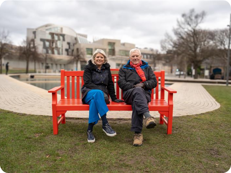 Sue and Jimmy Webber sit on a red bench in a park, a path is behind them