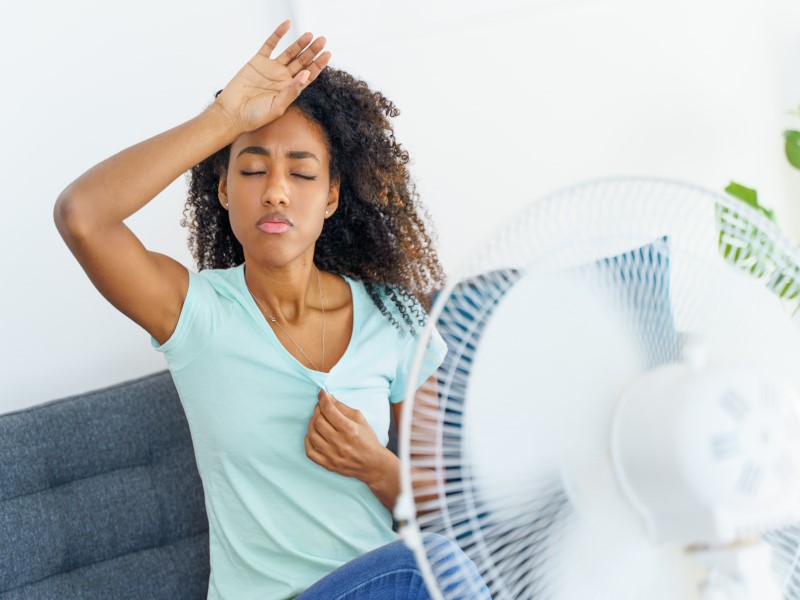 A young Black woman holds her hand to her forehead, looking visbly flushed. She is wearing a turquoise blue V neck t shirt. A white fan is pointing towards her with motion blur. 