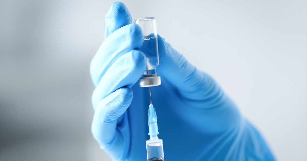 A gloved hand holds a small glass vial while a syringe draws liquid from it