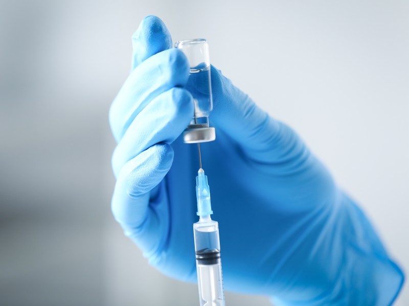 A gloved hand holds a small glass vial while a syringe draws liquid from it