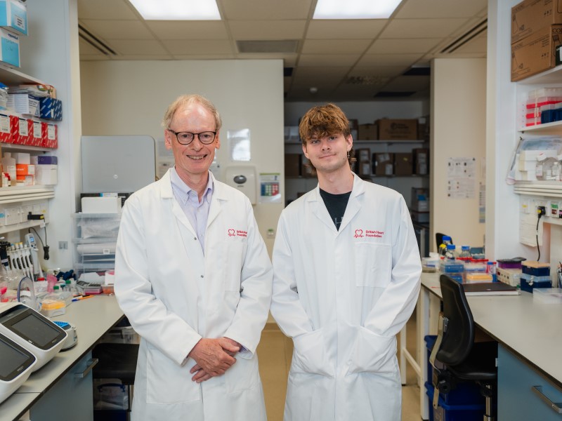 BHF Professor Hugh Watkins, an older white man with short hair, stands on the left in a laboratory next to Jack Taylor, a young white man with floppy brown hair. They are both wearing white lab coats which say British Heart Foundation in red writing on the right breast