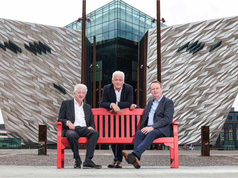 Two men sit either side of a red bench with a third man leaning over the bench behind them
