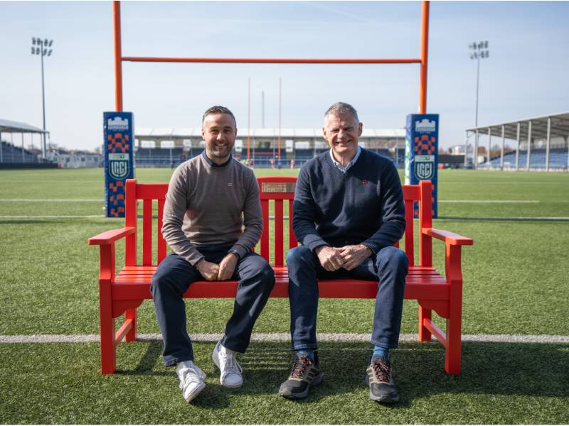 Two men sit on a red bench on a rugby pitch