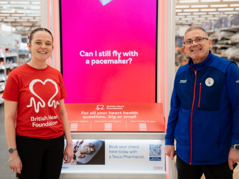 A White brown haired woman wearing a ponytail and bright red BHF T shirt is on the left, an older White man wearing glasses and with very short hair wearing a blue Tesco fleece stands on the right. You can't see their legs. In the middle is a red screen with the words 'Can I still fly with a pacemaker' in white writing. 