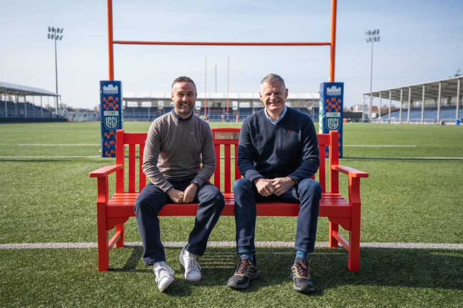 Two men sit on a red bench on a rugby pitch