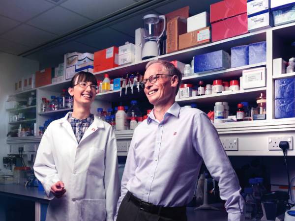a female researcher in a lab coat smiling and talking to a smartly dressed man as they stand in the office of a lab