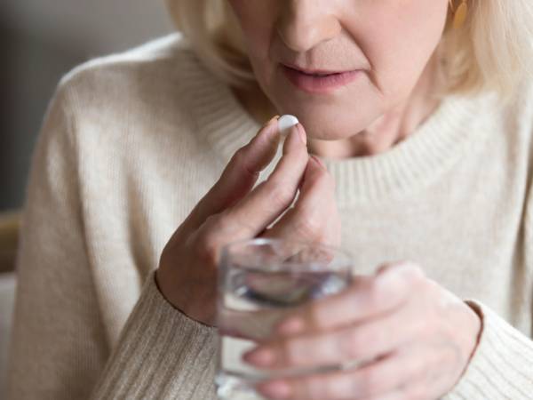 an older woman in a white jumper taking a pill with a glass of water